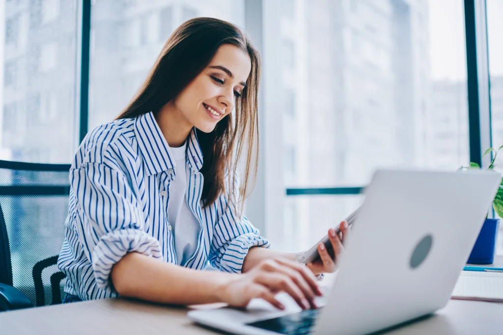 A woman is sitting in front of her laptop, holding a mobile phone